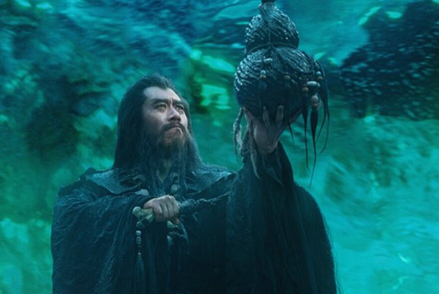 Still from a movie with a person wearing a cloak, with long hair and holding a mythical bird-like creature in his hand, against a blue/green background.
