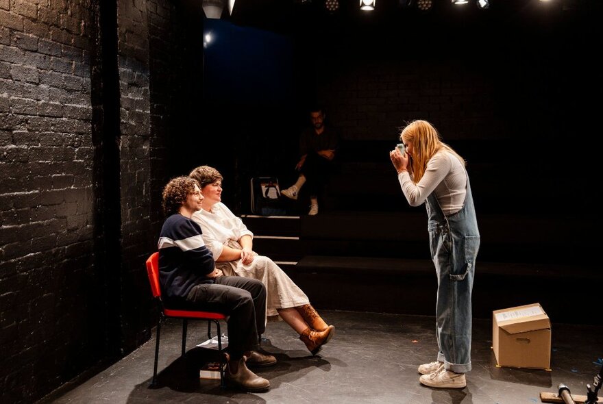 A man and a woman seated on red chairs on stage, with an 11-year old girl in overalls taking a photograph of them.