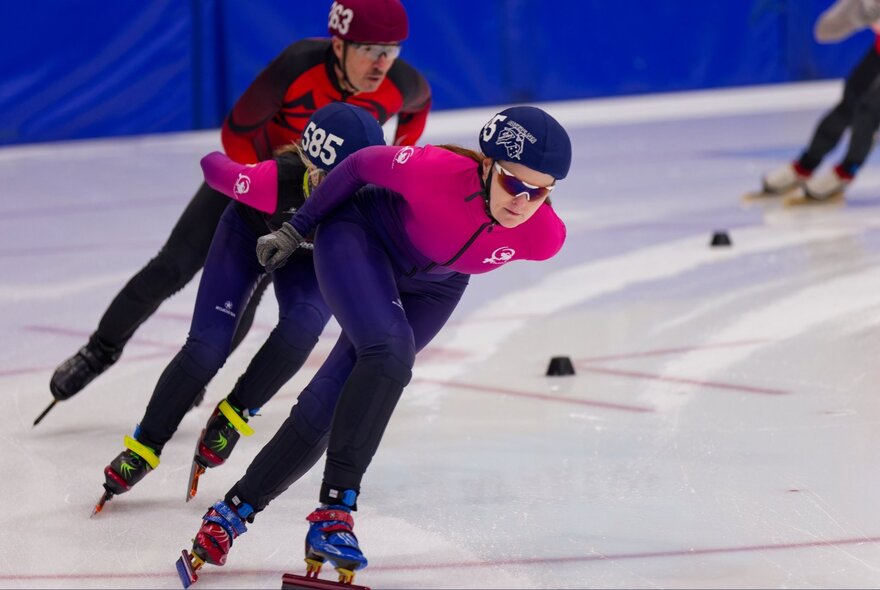 Short-track speed skaters rounding a corner on an indoor ice rink, wearing aerodynamic racing suits and protective helmets.