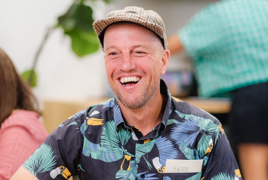 A man smiling openly with a bright shirt and tweed flat cap. 