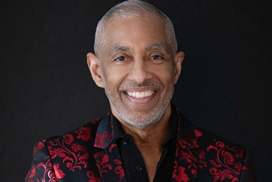 Smiling face of jazz musician Nestor Torres, wearing a patterned red and black shirt, posed against a black background.
