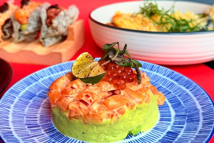 A Japanese dish with layers of green and pink garnished with lime and roe, on a blue patterned plate. 