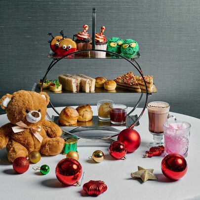 A children's themed three-tiered platter of high tea treats, alongside a teddy bear, drinks and Christmas baubles.