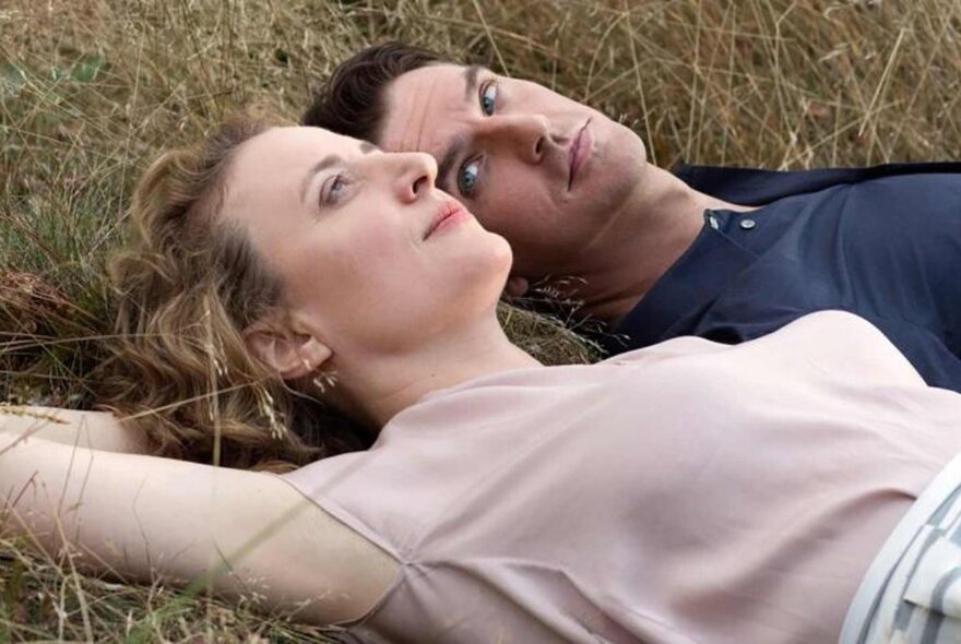 A man and a woman lying on the grass, the woman looking ahead, the man looking intensely at the woman.