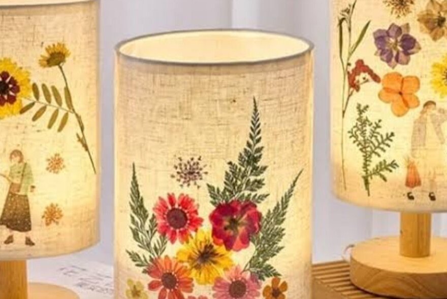 Three illuminated lamps with cylindrical parchment or fabric shades, decorated with various pressed flowers and small human figures. 