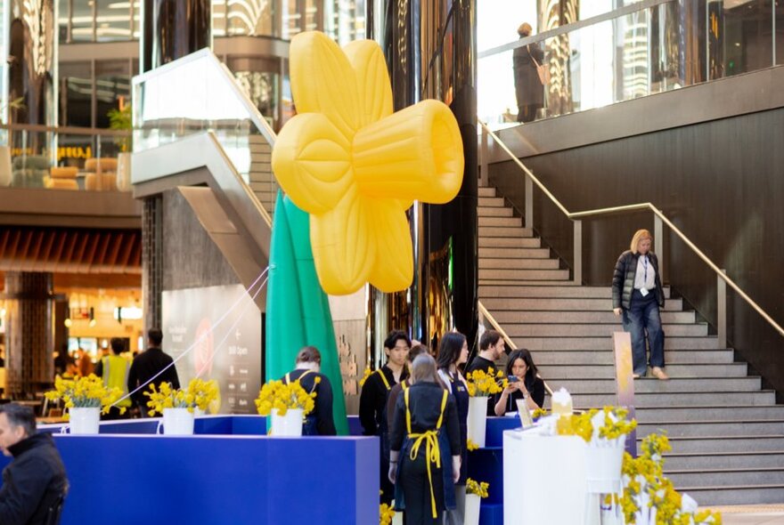 A 4m blow  up daffodil in the foyer of a shopping precinct, a sales counter with real daffodils surrounding it.