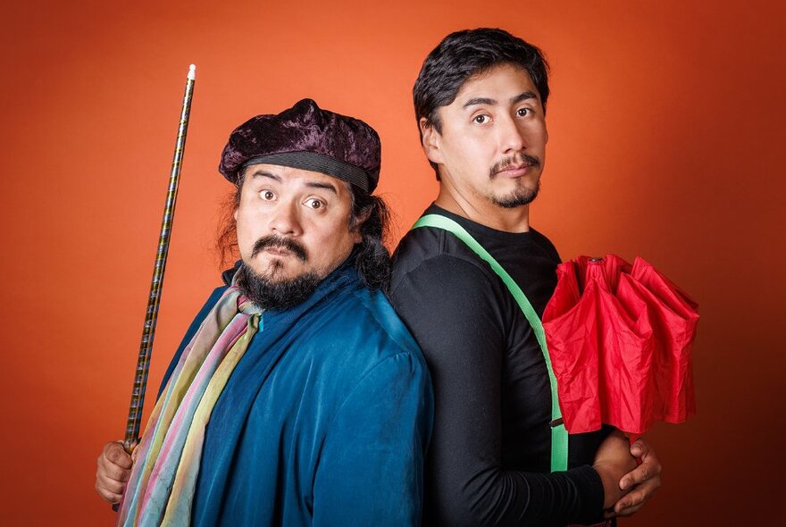 Two actors standing back to back in front of an orange backdrop, both looking at the viewer; one holding a long metal staff in his hand, the other a compact red umbrella.