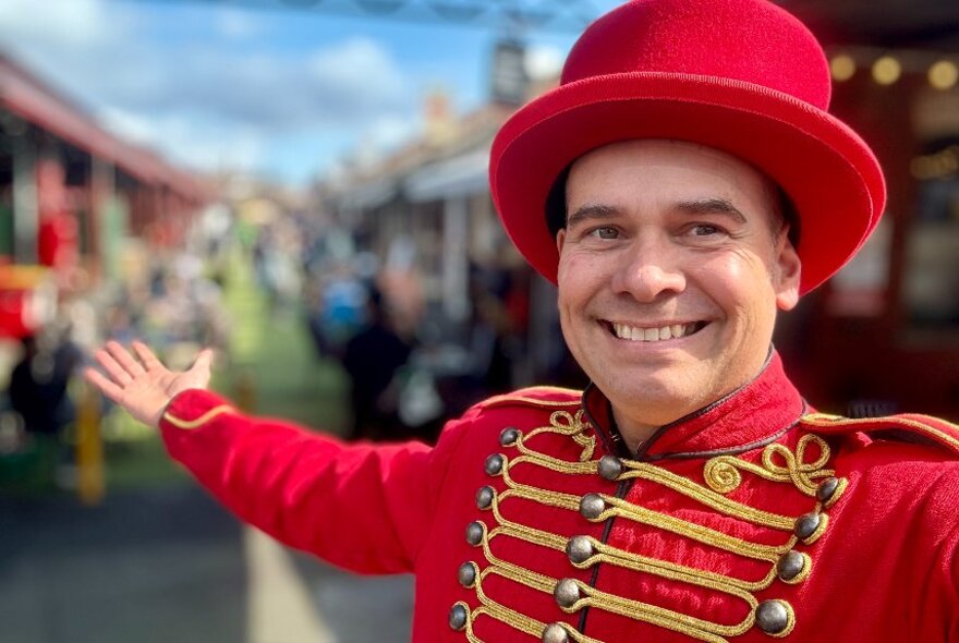 Christ Morant outdoors in front of a blurred background wearing a red ringmaster's coat and hat and holding out an arm.