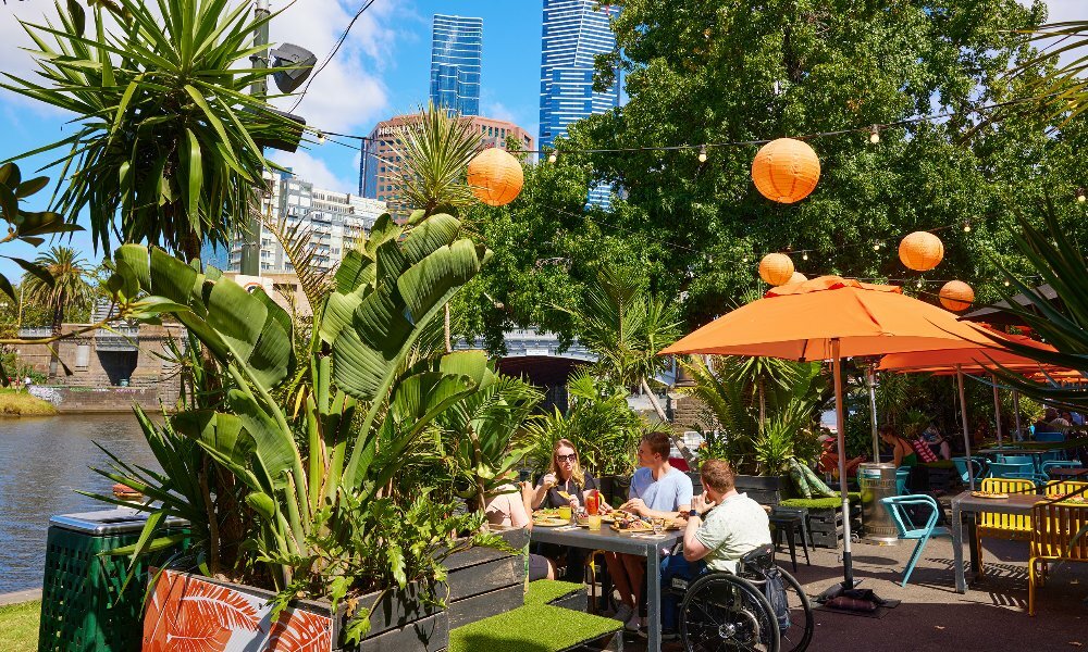 A group of friends are at a riverside bar drinking and eating