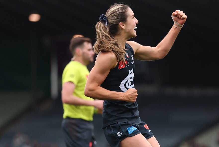 A Carlton AFLW player pumps her fist in celebration, a blurred umpire in the background. 