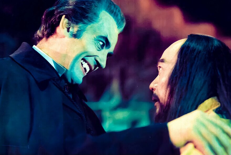 A still from the kung fu horror film, The Legend of the 7 Golden Vampires, of a vampire in profile, smiling at a man also in profile, who has long black hair. 
