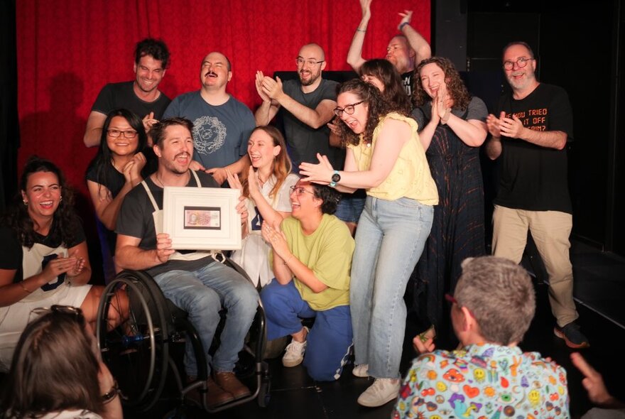 An ensemble cast of impro actors on a stage, all congratulating a person who is holding up a certificate.