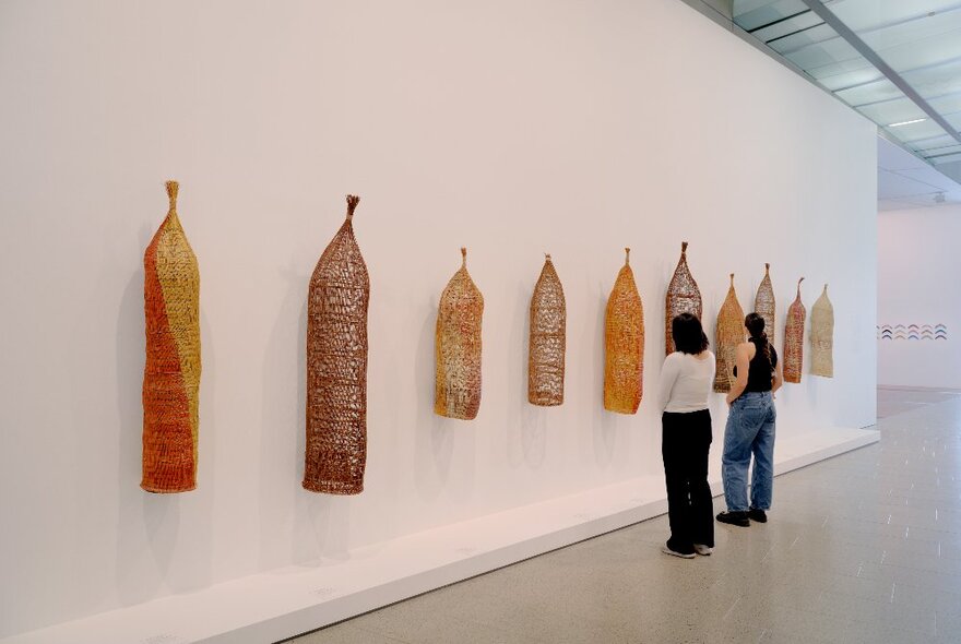 Long woven Indigenous artworks hanging in a gallery with two people looking on. 