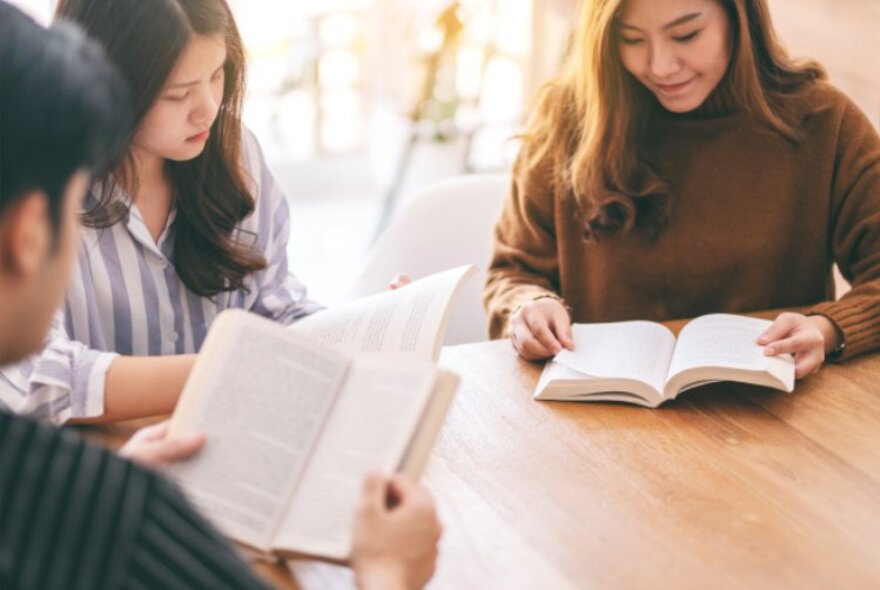 Three people seated around a wooden table, each reading a paperback novel.