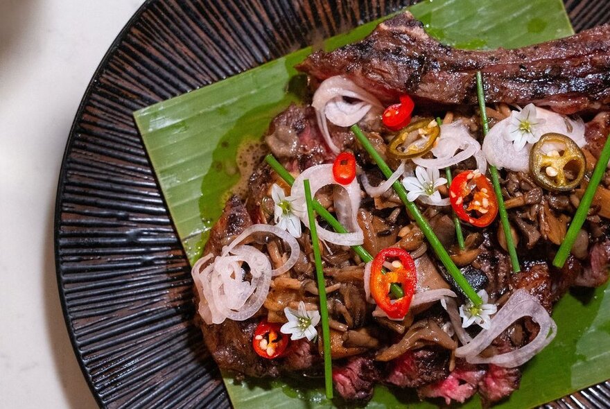 An Asian-inspired beef dish with chilli, onions and lemongrass stalks as garnish, presented on a banana leaf on a dark platter.
