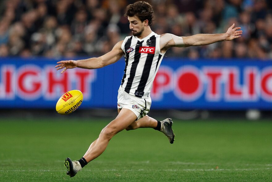 A Collingwood AFL player running on the field about to kick a yellow football, with a blurred stadium crowd behind him.