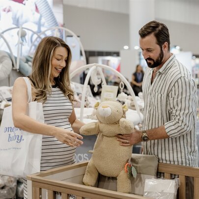 A man and pregnant woman hold a large teddy bear over a cot in a baby store. 