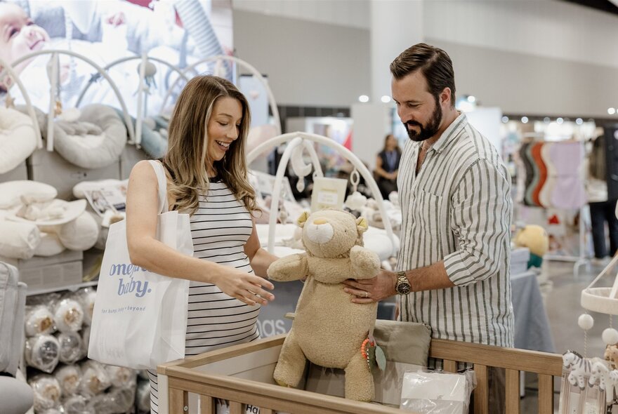 A man and pregnant woman hold a large teddy bear over a cot in a baby store. 