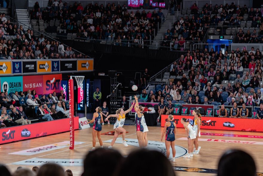 Netball teams, the Melbourne Vixens and the Sunshine Coast Lightening, play netball in an indoor stadium with a large crowd.