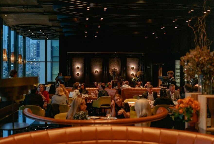 Patrons dining at a restaurant with curved leather booth seating and floor-to-ceiling windows.