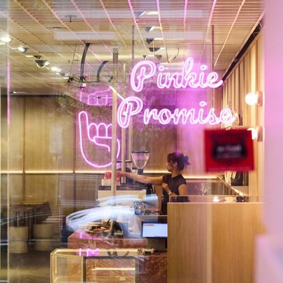 A barista working behind a coffee machine in a cafe with neon pink signage.