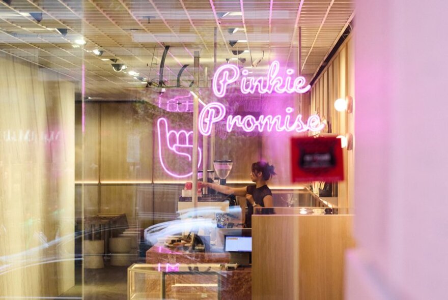 A barista working behind a coffee machine in a cafe with neon pink signage.