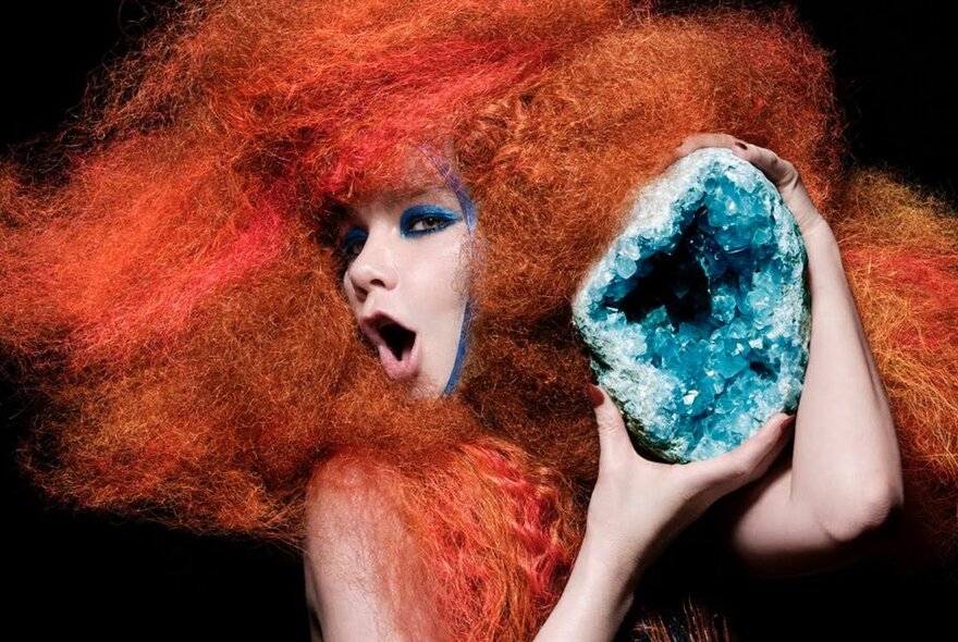 Artist Björk wearing a oversize red wig and holding a large blue crystal.