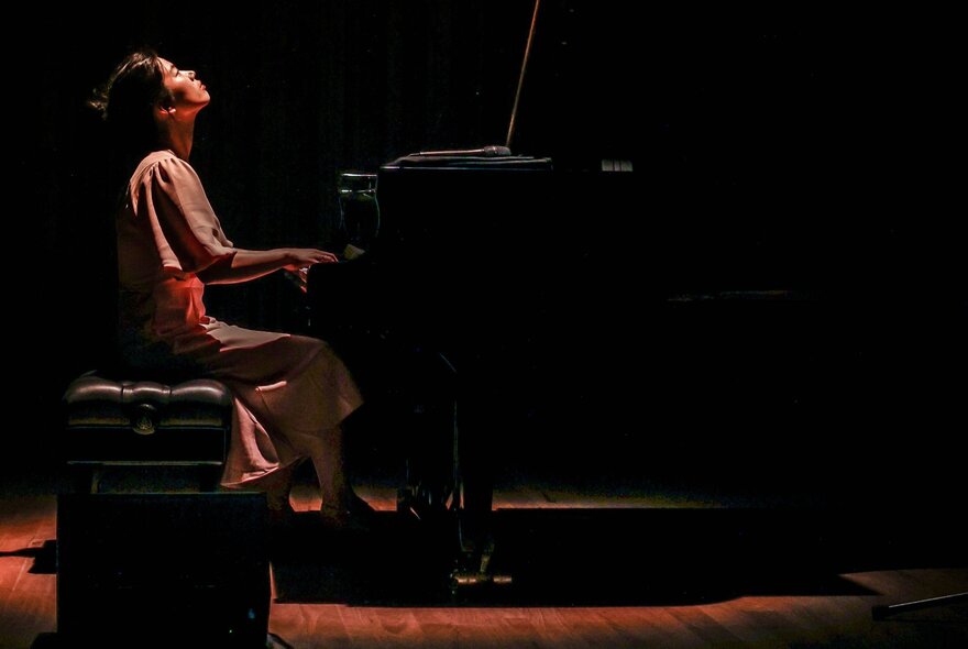 Pianist Mina Yu playing a grand piano while looking up at the ceiling, a single warm light cast down on her. 