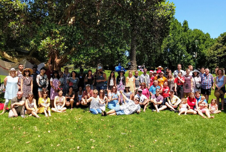 Large group of people sitting under the shade of a large tree in a park setting.
