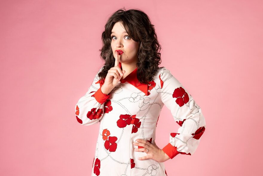 Singer and performer Alice Tovey in a white dress with a large red floral pattern on it, their finger in front of their lips in a 'Shh' gesture, standing in front of a pink backdrop.