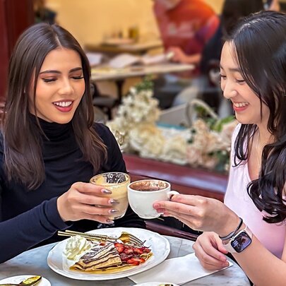 Two women holding coffees together in a celebratory cheers, with a crêpe to share on the round marble table between them.