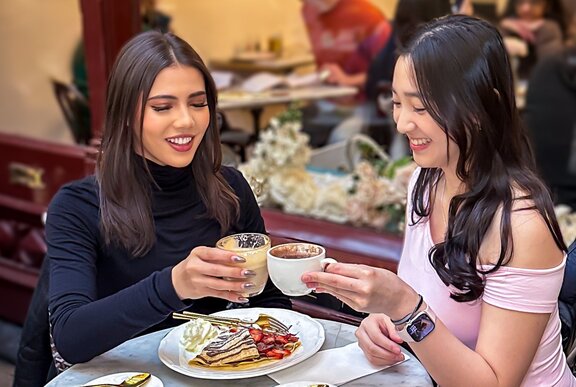 Two women holding coffees together in a celebratory cheers, with a crêpe to share on the round marble table between them.