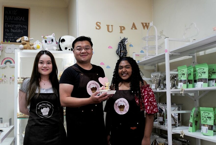 Three people standing in a group wearing branded aprons, the centre person is holding a cake featuring a small dog with a love heart.