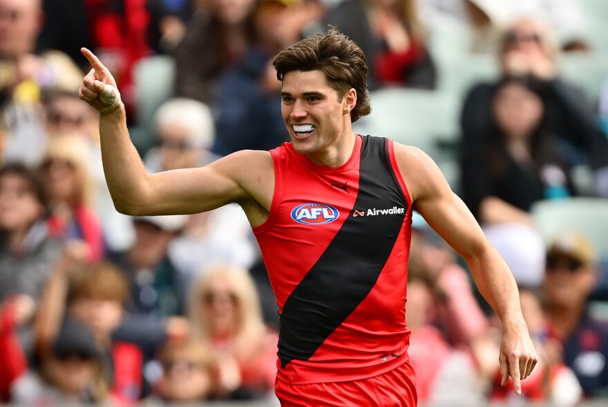 An Essendon AFL player celebrating on the field with one hand pointing, blurred footy fans in the stadium in the background. 