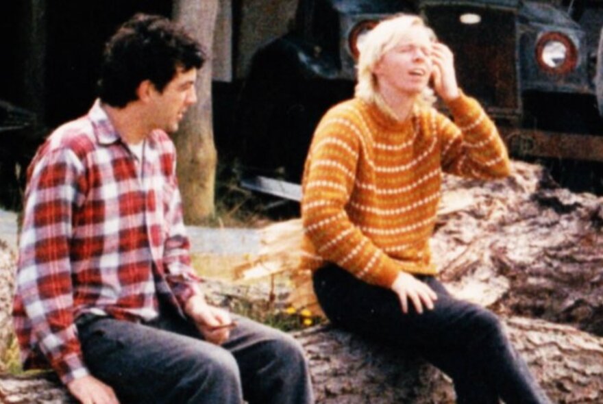 Vintage photo of two young men wearing plaid shirt and handknitted jumper, seated on a rock wall.