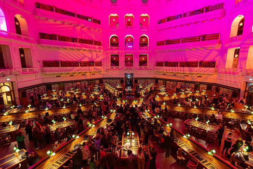 The Dome reading room of State Library Victoria showing the upper level balconies, and rows of desk fanning out from the centre, all bathed in a pink light.