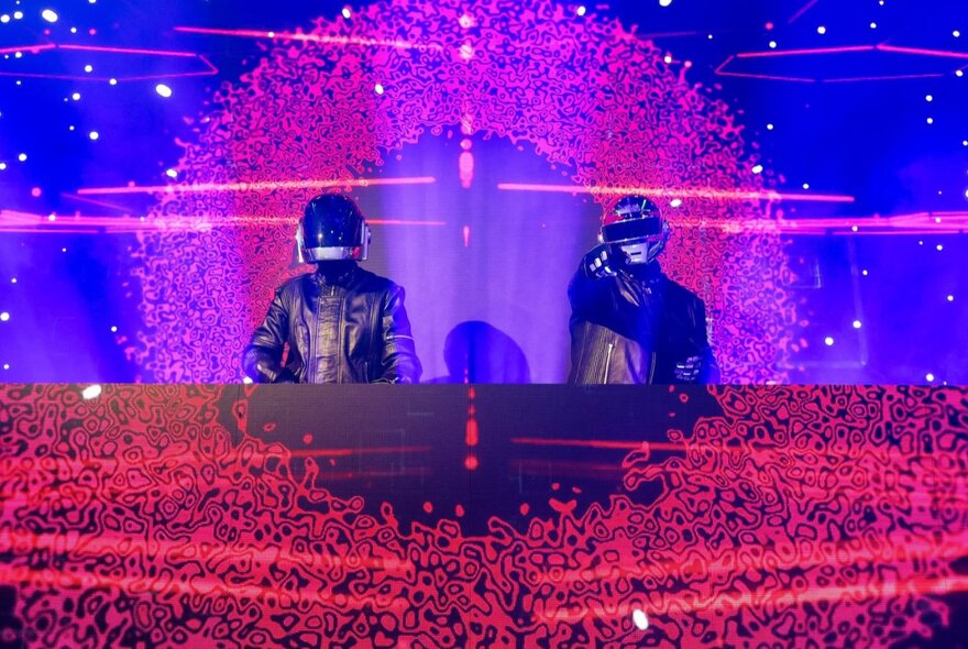Two DJs dressed in leather jackets and helmets like Daft Punk behind a neon-lit panel on stage.