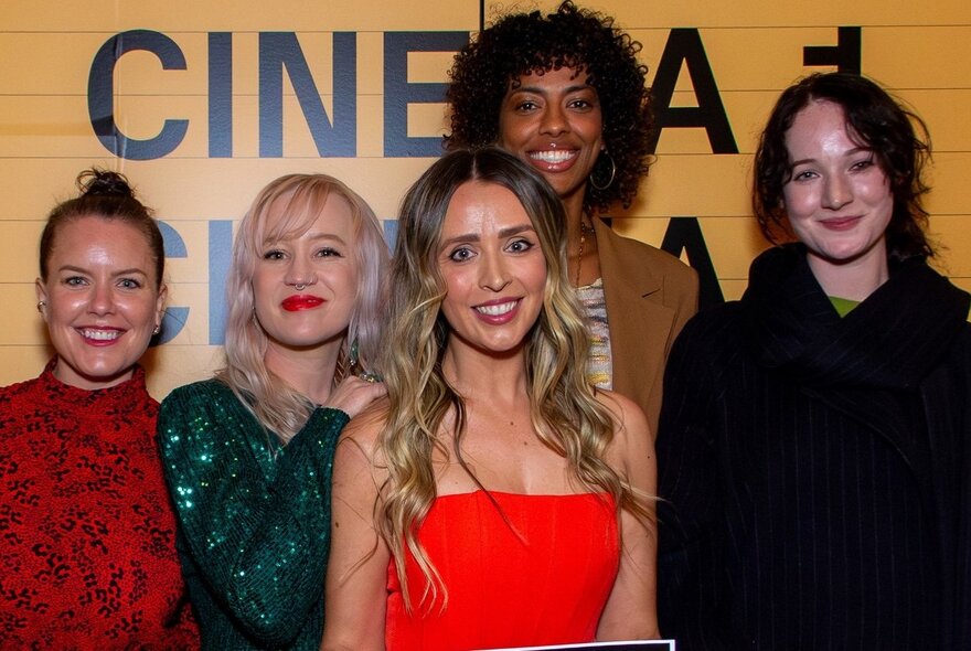 Five smiling people standing in front of a yellow wall that has the words CINEMA 1 in black letters partially visible. 