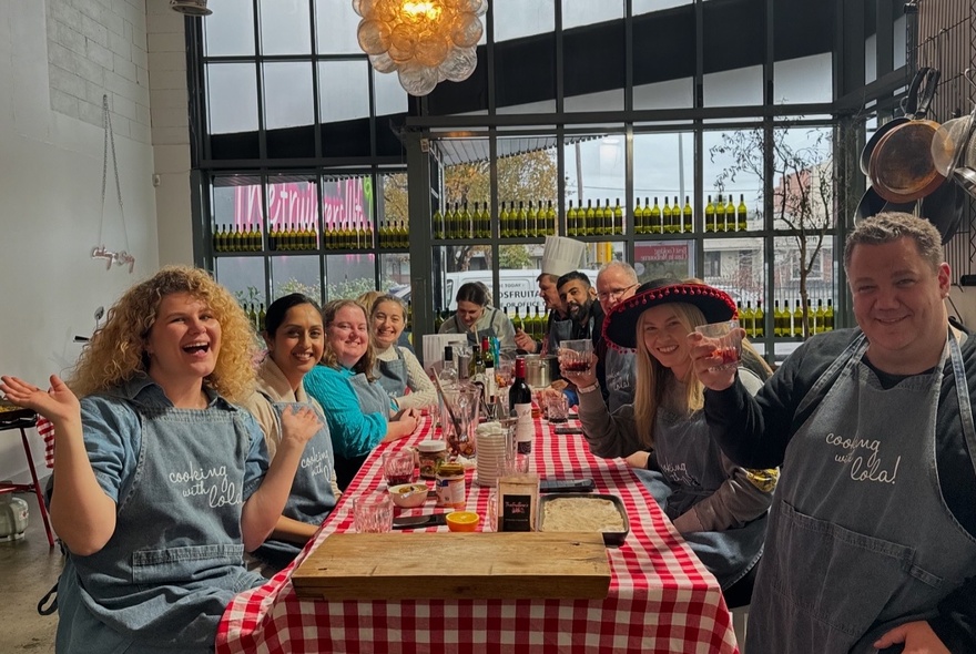 A group of people smiling and wearing aprons, seated at a long table participating in a cooking class.