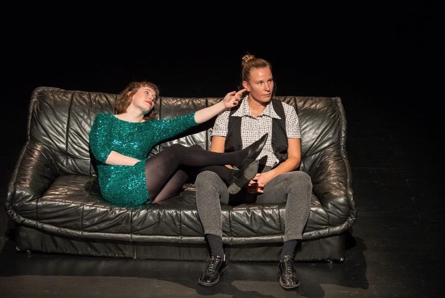 Two impro performers on a stage, sitting on a couch, one with their legs resting on the other's lap.