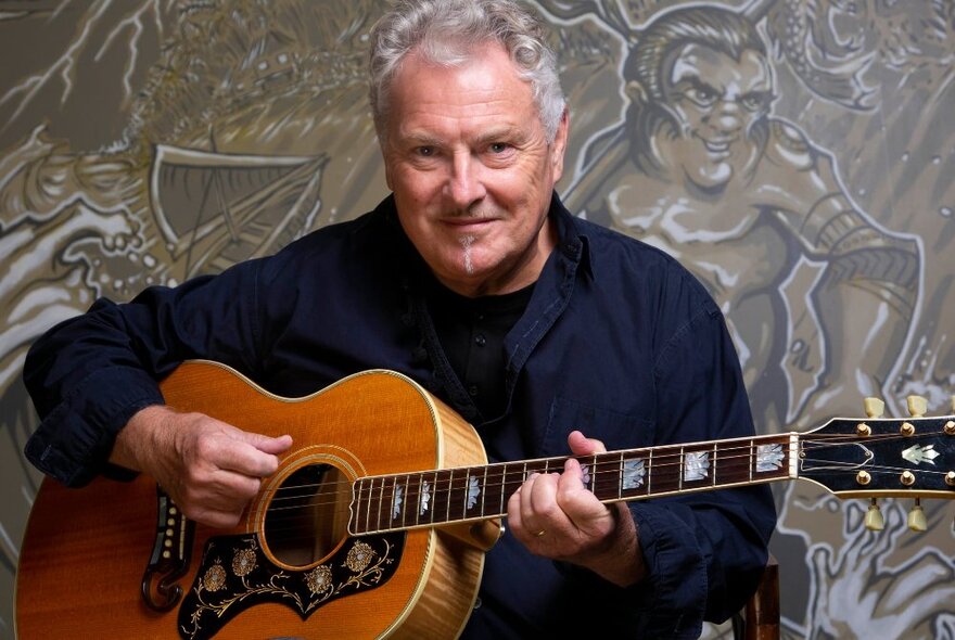 Singer and musician Ross Wilson holding an acoustic guitar and smiling.