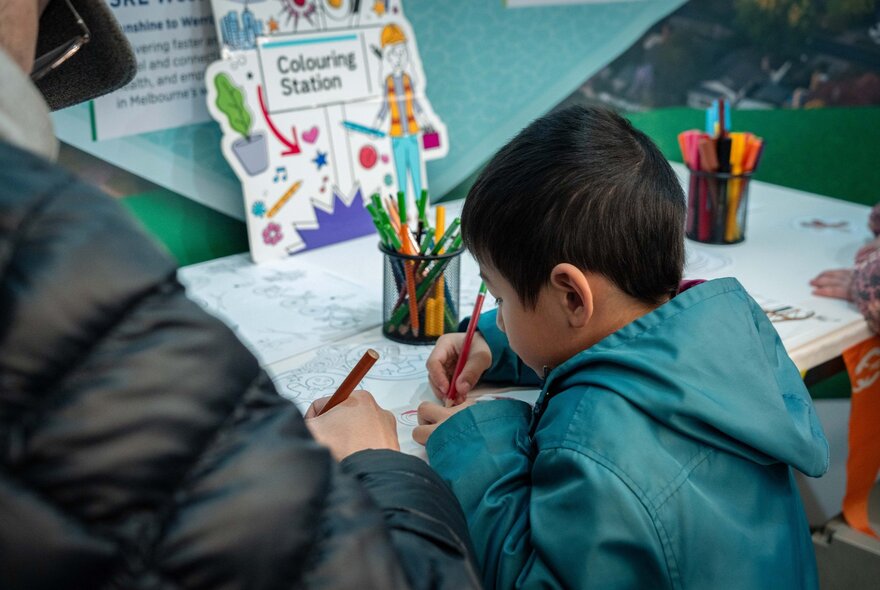 A young child at a craft table, a jar of coloured pencils in front of them, colouring in.