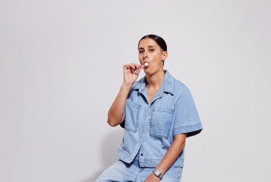 Comedian Annie Boyle seated on a stool wearing a denim shirt and denim shorts, sucking on a Chupa Chups lollipop, posed against a white background.