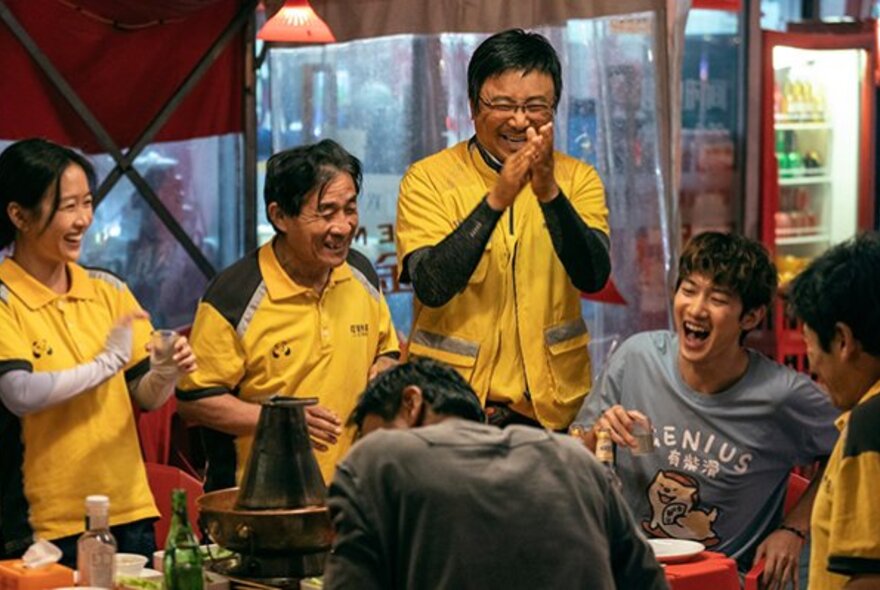 Still from a movie depicting a group of people, some of them wearing the same yellow and black uniform style shirt, laughing and socialising around a table.