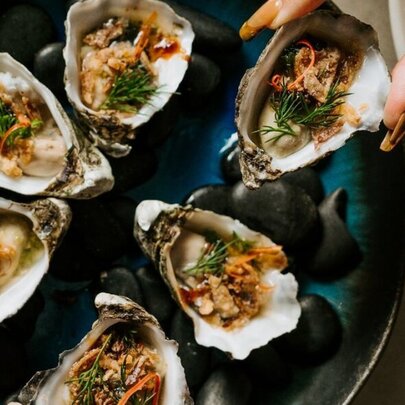 A hand delicately holding an opened oyster above a plate of oysters.