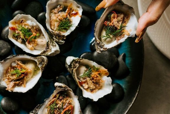 A hand delicately holding an opened oyster above a plate of oysters.