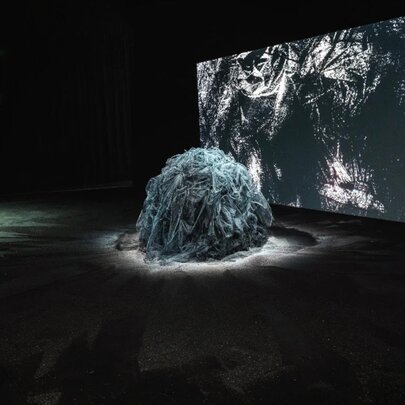 A large, mounded form made from strips of fabric, spotlit in front of a large screen in an exhibition space.