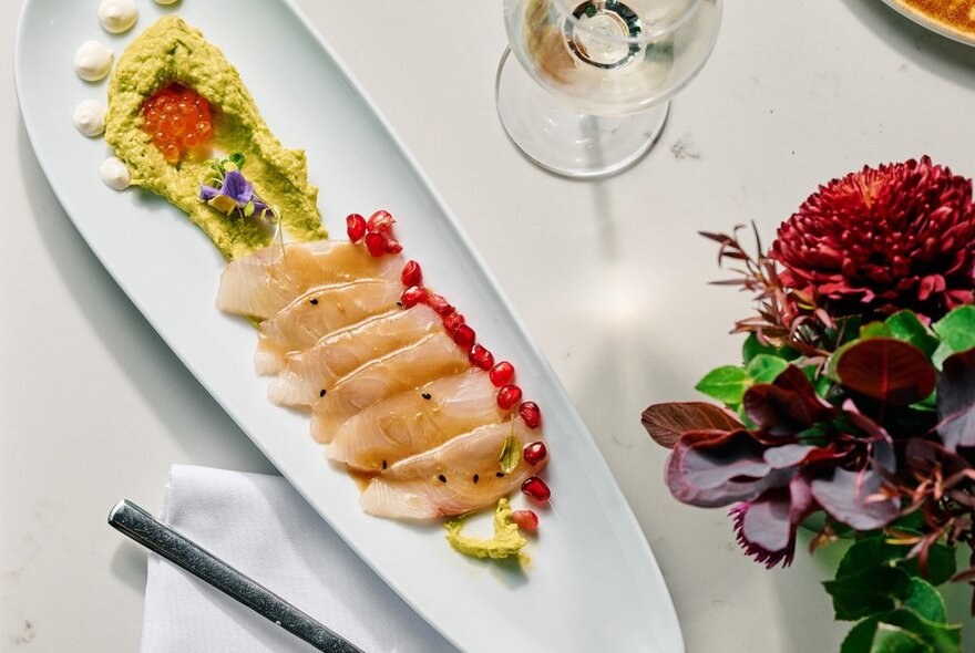 A long white platter of seafood with garnish, flowers and glass of wine.