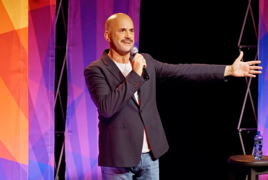 Costa Carastavrakis, a bald man on stage talking into a microphone, wearing a navy suit jacket and jeans.