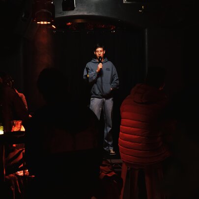 A comedian holding a microphone on stage in a darkened venue.